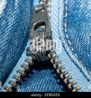 Makroaufnahme eines Metallreißverschlusses auf blauem Denim-Gewebe mit Reißverschlusszähnen, Schieberegler und Textil-Textur mit geringer Schärfentiefe. Stockfoto