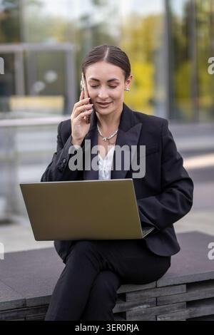 Eine Frau sitzt draußen an einem Tisch. Sie trägt einen schwarzen Anzug und spricht mit ihrem Telefon. Ihr Laptop ist vor ihr offen, als sie verlobt aussieht Stockfoto