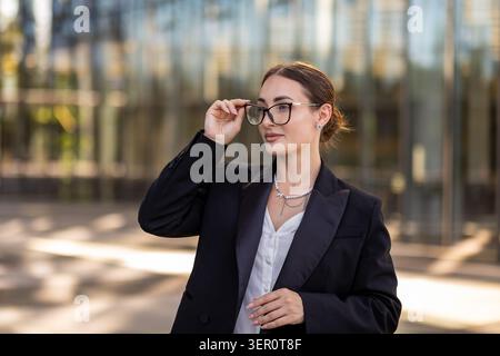 Eine Frau steht draußen vor einem Glasgebäude. Sie trägt einen dunklen Blazer und passt ihre Brille an, während sie wegblickt. Es ist ein heller Tag wi Stockfoto
