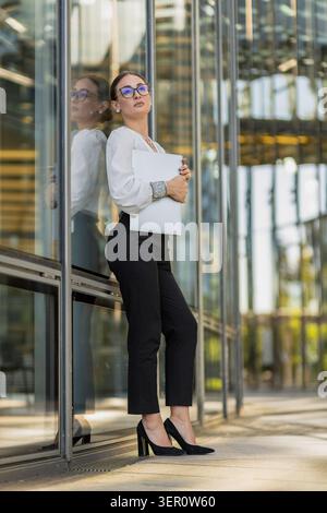 Eine Frau in formellem Outfit steht vor einem verglasten Bürogebäude. Sie hält eine Mappe in der Hand und schaut selbstbewusst in die Kamera. Das Sonnenlicht scheint Stockfoto