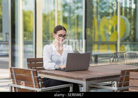 Eine Frau sitzt an einem Holztisch vor einem Glasgebäude und konzentriert sich auf ihren Laptop. Sie tippt, während sie eine Brille trägt. Sonnenlicht hebt die Szene hervor, kreativ Stockfoto