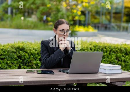Eine Frau sitzt an einem Holztisch im Park und konzentriert sich auf ihren Laptop. Sie hat ihre Hände gefesselt und trägt eine Brille. In der Nähe gibt es ein Telefon, ein Tablet und Stockfoto