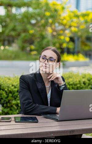Eine Frau sitzt an einem Tisch im Park und arbeitet an ihrem Laptop. Sie sieht nachdenklich aus, während sie eine Brille und einen schwarzen Anzug trägt. Zwei Geräte sind neben ihr, an Stockfoto