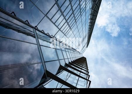 Die Leute laufen auf der Straße, während sie auf ein hohes Glasgebäude schauen. Die Struktur reflektiert den Himmel und die Wolken und bietet einen beeindruckenden Blick von unten. Stockfoto