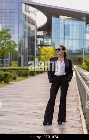 Eine Frau steht auf einem Weg in einem Geschäftsviertel. Sie trägt einen schwarzen Anzug und eine Sonnenbrille. Die Sonne scheint auf die modernen Gebäude um sie herum. Stockfoto