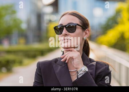 Eine Frau steht selbstbewusst in einem Stadtgebiet, trägt eine Sonnenbrille und eine schwarze Jacke. Sie hat ihre Hand am Kinn, blickt nach vorne, mit grünen Bäumen und Stockfoto