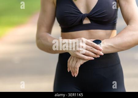 Eine Frau ist draußen in einem Park. Sie trägt Trainingskleidung und überprüft ihren Aktivitätstracker am Handgelenk. Die Sonne ist draußen und der Weg ist sichtbar Stockfoto