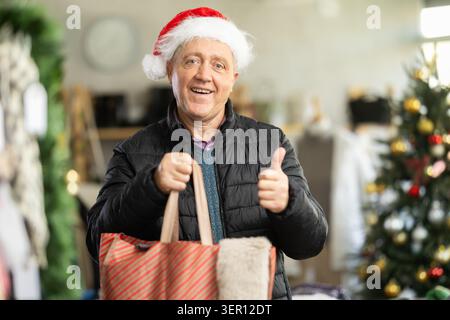 Ein reifer Mann hält in einem Laden eine ganze Tüte voller Dinge vor dem Hintergrund eines Weihnachtsbaumes Stockfoto