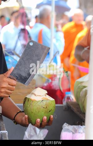 Nahaufnahme von Händen, die frische junge Kokosnüsse mit einem Beil auf einem thailändischen Straßenmarkt öffnen, mit verschwommenen Leuten im Hintergrund. Stockfoto