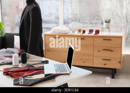 Laptop mit Nähzubehör und Herrenkleidung auf Schneidertisch im Atelier Stockfoto