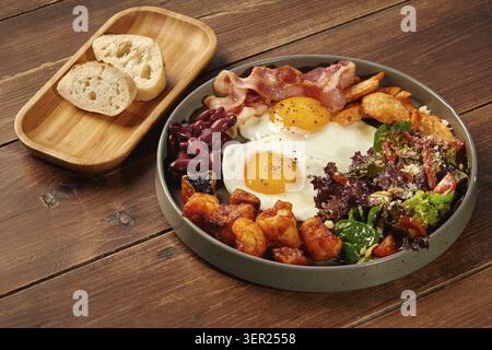 Grauer Teller mit einem kompletten Frühstück aus Spiegeleiern, Speck, Bohnen, Hühnchen, Kartoffeln und Salat, serviert auf einem Holztisch mit Brot Stockfoto