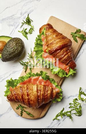 Zwei frisch gebackene Croissant-Sandwiches gefüllt mit Räucherlachs, Salat und Avocado, serviert auf einem Holzbrett Stockfoto
