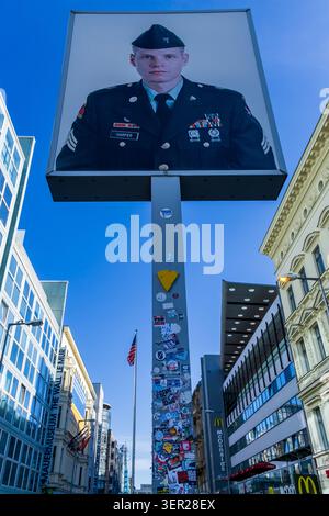 Tiefwinkelansicht des ikonischen US-Soldaten-Porträts am Checkpoint Charlie in Berlin, Deutschland, mit dem Mauermuseum, der amerikanischen Flagge und dem modernen Stadtbild. Stockfoto