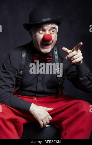 Porträt eines Clown. Ein älterer Mann mit roter Clownnnase und Bowler-Hut auf dem Kopf. In roten Hosen und einem schwarzen Hemd mit Hosenträgern gekleidet. Der Clown poin Stockfoto