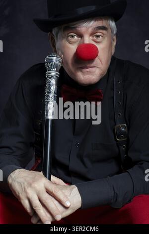 Dramatisches Porträt eines traurigen Clowns vor dunklem Hintergrund. Ein älterer Mann mit roter Clownnnase, roter Hose und Bowler-Hut auf dem Kopf. In schwarzem Shi gekleidet Stockfoto