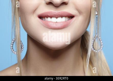 Makroporträt des weiblichen unteren Gesichtsbereichs. Blonge Frau mit roten, lächelnden Lippen, Make-up und Ohrringen. Mädchen auf blauem Hintergrund Stockfoto