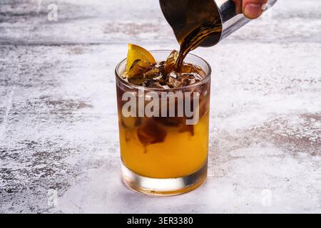 Dunkler Espresso von Hand in Glas gefüllt mit Orangensaft und Eiswürfeln, garniert mit Orangenscheibe, auf rustikalem weißem Holzhintergrund, r Stockfoto