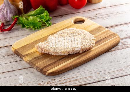 Rohes paniertes Hähnchenkotelett auf rustikalem hölzernem Schneidebrett mit frischem Salat Knoblauch und roter Paprika im Hintergrund. Lebensmittelzubereitungskonzept mit Stockfoto