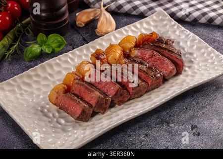 Köstlich gebratenes, in Scheiben geschnittenes picanha-Steak, mittelselten, präsentiert auf strukturiertem weißen rechteckigen Teller. Garniert mit frischem grünem Basilikum und roter Kirschtoma Stockfoto