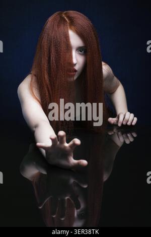 Schöne erschreckende junge Frau mit Ingwerhaar über Spiegelspiegel auf blauem Hintergrund Stockfoto