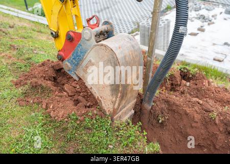 Nahaufnahme eines Mini-Baggerlöffels aus Metall, der in braunen Boden direkt neben einem schwarzen flexiblen Versorgungsschutzrohr oder einer Kabelhülse gräbt. Hoch Stockfoto
