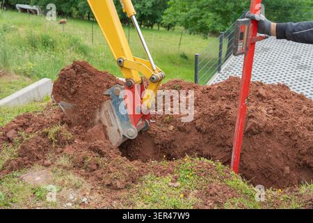 Ein gelber Mini-Baggerlöffel, der tief in braunen Boden gegraben wird, neben einem roten Vermessungsstab für die Landschaftsgestaltung im Garten. Professionelle Bodenvorbereitung Stockfoto