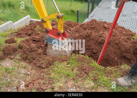 Nahaufnahme eines hydraulischen Minibaggerlöffels, der dunkelbraune Erde auf einem Wohngrundstück gräbt, während ein Arbeiter in der Nähe steht. Professionell Stockfoto