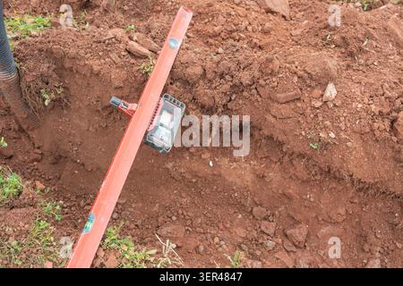 Ein professionelles orangefarbenes Wasserwaage-Messgerät mit digitalem Messempfänger, das in einem Schmutzgraben platziert ist, um die Genauigkeit der Neigung zu prüfen Stockfoto