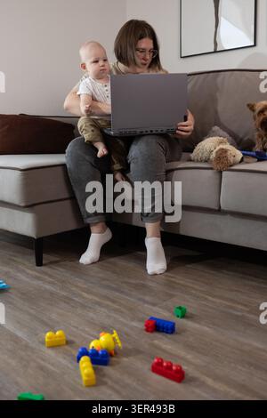 Ein sitzender Elternteil hält ein Kind auf dem Schoß, während er auf einem Sofa in neutraler Farbgebung im Wohnzimmer an einem Laptop arbeitet. Bunte Plastikbausteine und ein kleines Stockfoto