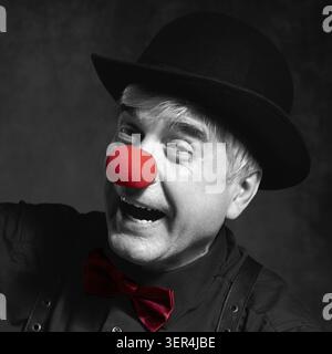 Selektives Farbporträt eines lachenden Clowns vor dunklem Hintergrund. Ein älterer Mann mit roter Clownnnase und Bowler-Hut auf dem Kopf. In schwarzem Shir gekleidet Stockfoto
