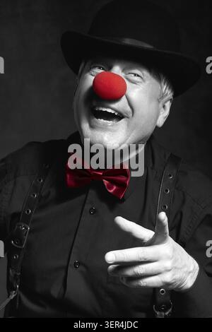 Selektives Farbporträt eines lachenden Clowns vor dunklem Hintergrund. Ein älterer Mann mit roter Clownnnase und Bowler-Hut auf dem Kopf. In schwarzem Shir gekleidet Stockfoto