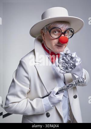 Ein älterer Clown in einem weißen Mantel und Hut mit Gehstock und einer Clownmedaille in der Hand Stockfoto