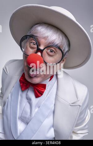 Ein älterer Clown mit weißem Mantel und Hut. Der Clown, der seine Zunge rausstreckt, mit einem schlauen Ausdruck im Gesicht Stockfoto