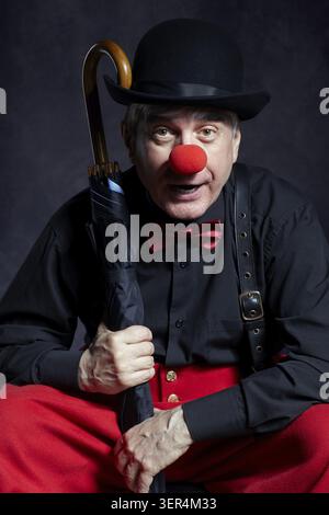 Porträt eines Clown mit Regenschirm auf dunklem Hintergrund. Ein älterer Mann mit roter Clownnnase und Bowler-Hut auf dem Kopf. In roten Hosen und schwarzem Hemd gekleidet Stockfoto