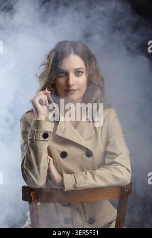 Porträt einer jungen, schönen blonden Frau sitzt auf einem Stuhl. Weiblich in beigem Mantel auf rauchem Hintergrund Stockfoto