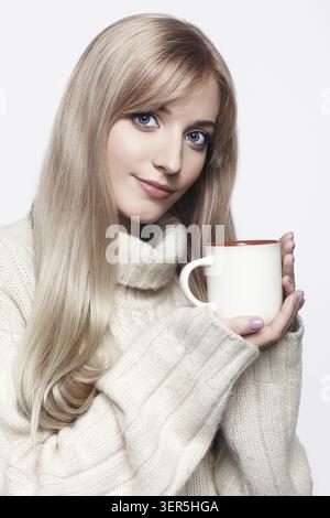 Blonde junge Frau mit einer Kappe Tee in den Händen. Frau in einem großen weißen Kaschmirpullover, der auf grauem Hintergrund posiert Stockfoto