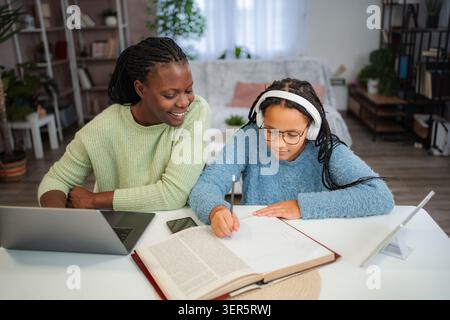 Eine Mutter und ihre Tochter verbringen einen warmen Moment in ihrem Heimbüro, während sie sich auf das Lernen konzentrieren. Die Mutter lächelt, während ihre Tochter in einem Notizbuch schreibt Stockfoto