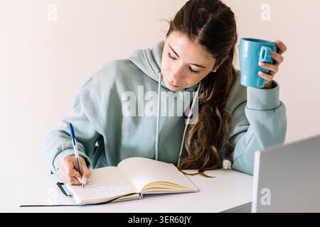 Eine Frau sitzt an einem Schreibtisch mit einem blauen Becher in der Hand. Sie schreibt in ein Notizbuch Stockfoto