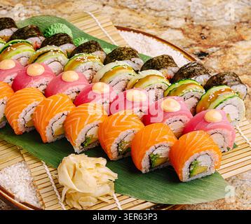 Eine wunderschöne Präsentation von Sushi-Brötchen in verschiedenen Farben ist auf einer Bambusplatte angeordnet. Das Sushi umfasst Lachs, Thunfisch, Avocado und Gurke mit Ingwer- und Wasabi-Garnituren. Stockfoto
