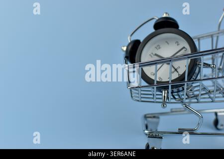 Mini-Uhr sitzt im Warenkorb. Metallwagen für Uhr mit schwarzem Rahmen. Weißes Zifferblatt, schwarze Zeiger, einfache Ziffern. Hellblauer Hintergrund Stockfoto