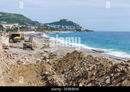 Bautätigkeit an einem Strand mit schweren Maschinen und Sandhaufen, mit einer Küstenstadt und Bergen im Hintergrund unter klarem Himmel Stockfoto