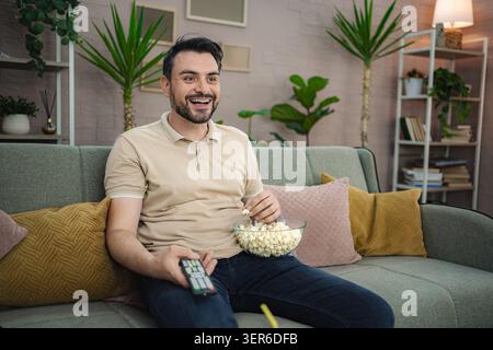 Mann lacht, während er fernsehen und Popcorn auf dem Sofa zu Hause isst Stockfoto