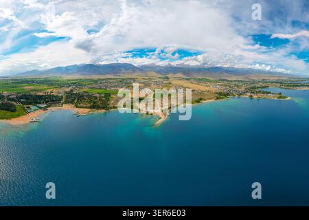 Aus der Vogelperspektive fängt der riesige blaue Issyk-Kul-See ein, der von Bergen und grünen Feldern umgeben ist. Die malerische Küstenlinie umfasst Strand- und Resortgebäude unter Ccle Stockfoto