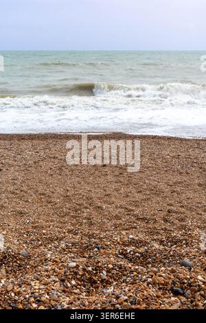 Die Flut rollt ein und aus, während die Wellen auf einen Kieselstrand stürzen. Der Strand erstreckt sich entlang der Küste unter bewölktem Himmel. Kieselsteine bedecken den Sand während Stockfoto