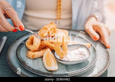 Eine Frau, die eine Gabel benutzt, um einen knusprigen, gebratenen Calamari-Ring zu essen und ihn in eine kleine Schüssel Sauce zu tauchen. Zitronenkeil auf Teller Stockfoto