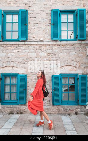 Frau, die in einem roten Kleid vor einer Steinmauer mit leuchtenden blauen Fensterläden steht und Architektur und Straßenstil erforscht Stockfoto