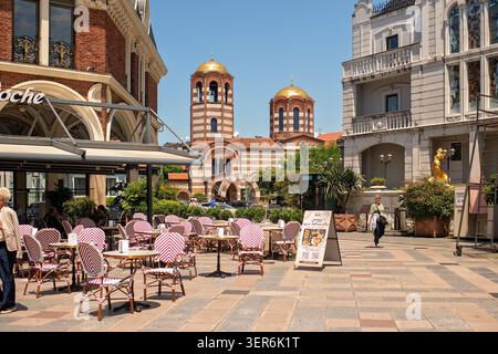 Batumi, Georgia - 9. Juni 2026: Straßencafé-Terrasse mit gestreiften Korbstühlen und goldenen Kuppeln der St. Nikolaus-Kirche auf dem Piazza Square. Stockfoto