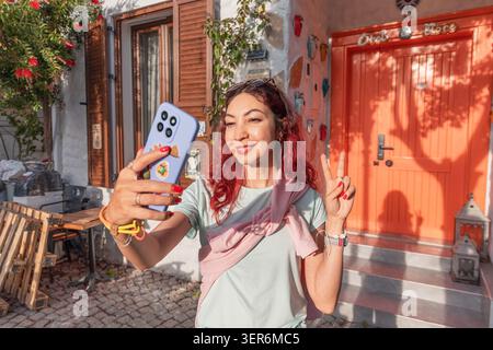 13. Mai 2026, Sigacik, Turkiye: Frau mit roten Haaren lächelnd, Telefon für ein Selfie mit einem Friedensschild vor einer orangen Tür, unterwegs Stockfoto