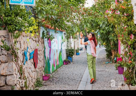13. Mai 2026, Sigacik, Turkiye: Junge Frau, die entlang einer malerischen Kopfsteinpflasterstraße mit blühenden Pflanzen, Steinmauern und lebendiger Wandkunst spaziert Stockfoto