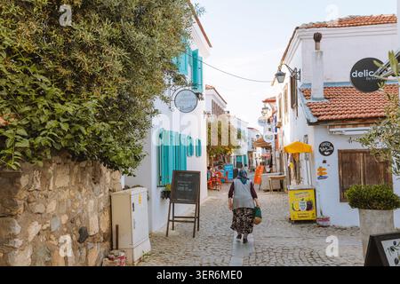 13. Mai 2026, Sigacik, Turkiye: Frau, die entlang einer traditionellen kopfsteingepflasterten Gasse mit weißen Häusern und türkisfarbenen Rollläden spaziert Stockfoto